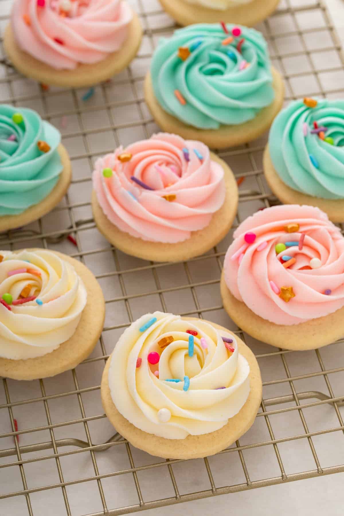 Sugar cookie frosting piped into rosettes in three different colors  and topped with rainbow sprinkles on round sugar cookies that are resting on a wire rack.
