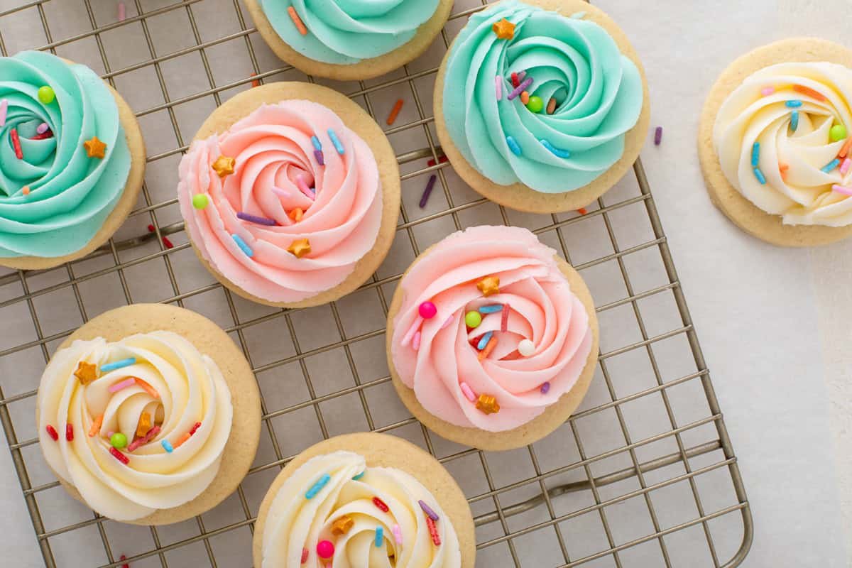 Sugar cookie frosting piped into rosettes in white, pink, and mint green and topped with rainbow sprinkles on round sugar cookies.