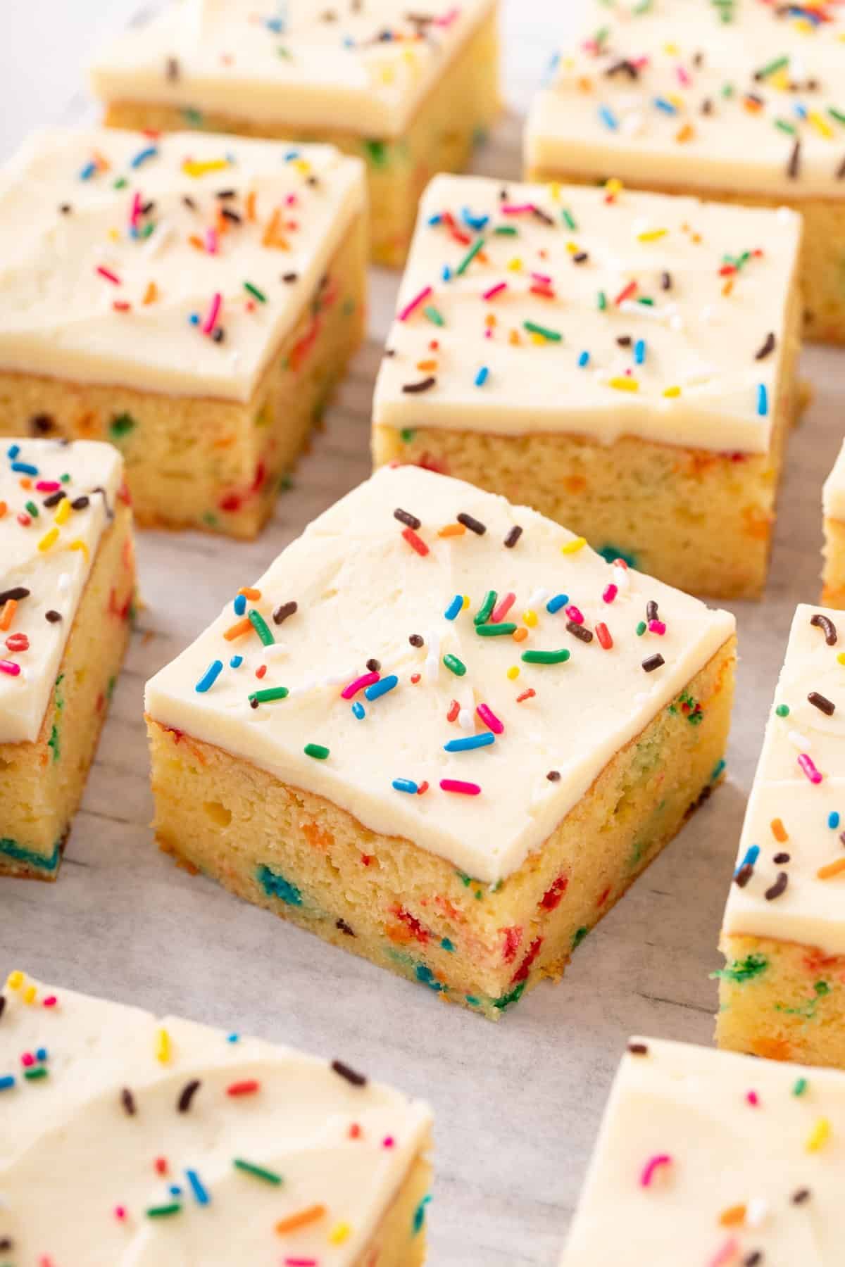Sliced funfetti sheet cake frosted with vanilla frosting and topped with rainbow sprinkles.