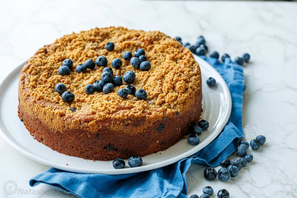 whole round blueberry coffee cake on a white dish with blueberry garnish and blue tea towel