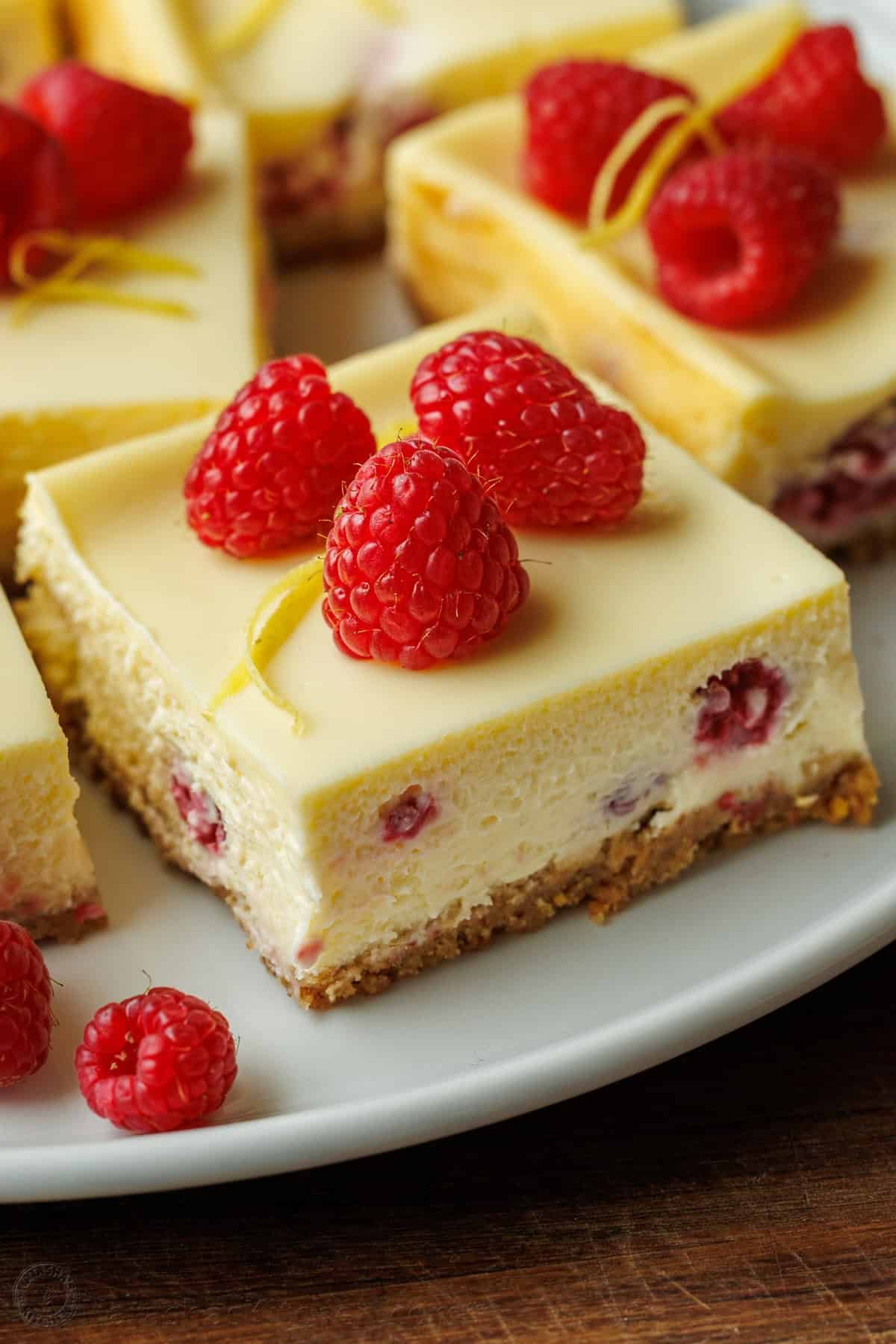 One raspberry lemon cheesecake bar on a plate with raspberries and lemon peel garnish