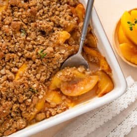 Spoon about to scoop peach crisp out of a white baking dish.