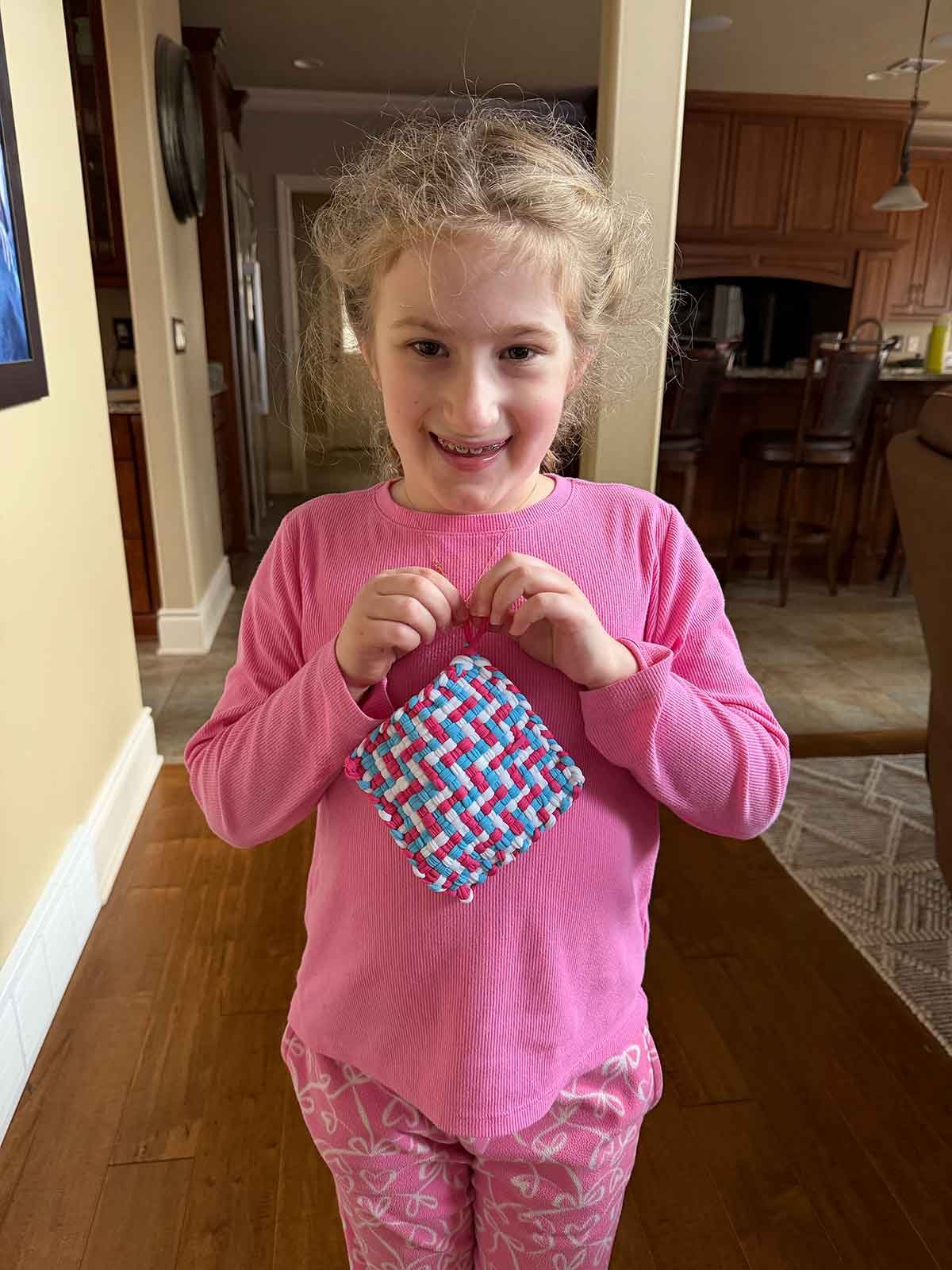Girl in pink pajama set holding a pot holder made with a weaving loom.