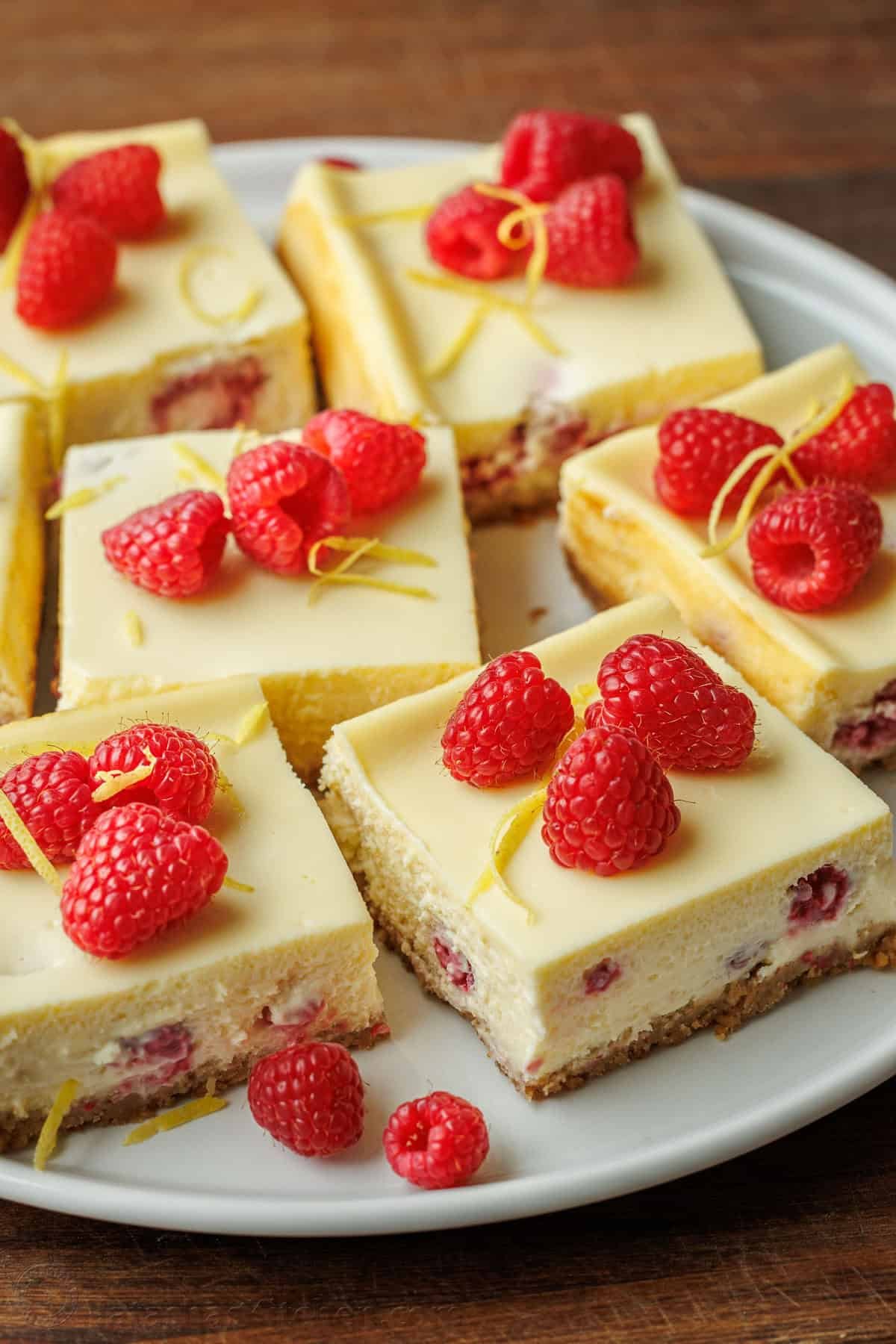 Raspberry Lemon Cheesecake Bars on a white serving platter with raspberries and lemon zest