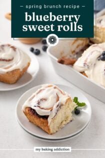 White plate holding a blueberry sweet roll, with a second plate and the pan of rolls in the background. Text overlay includes recipe name.