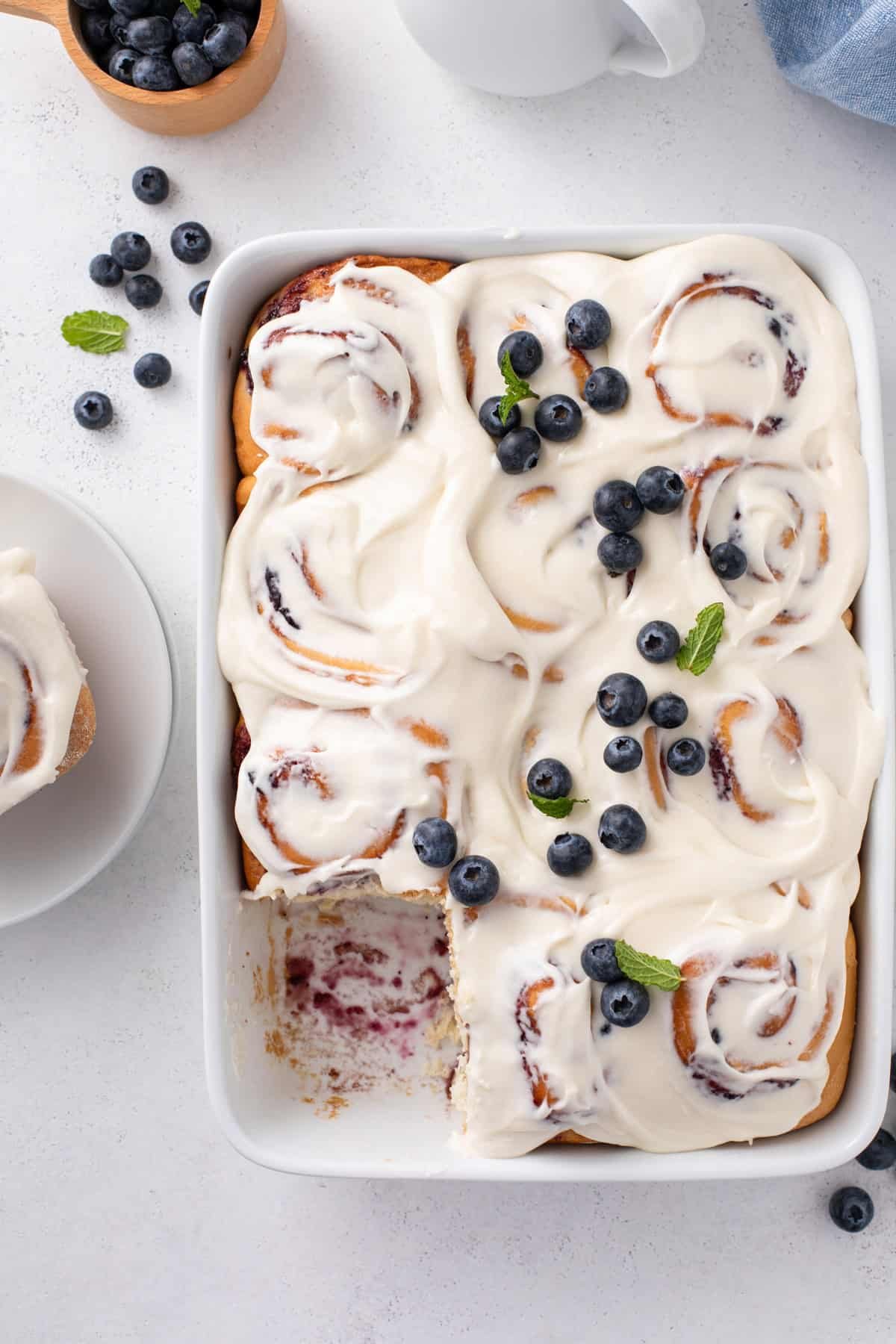 Overhead view of a pan of blueberry sweet rolls with one roll removed from the corner of the pan.