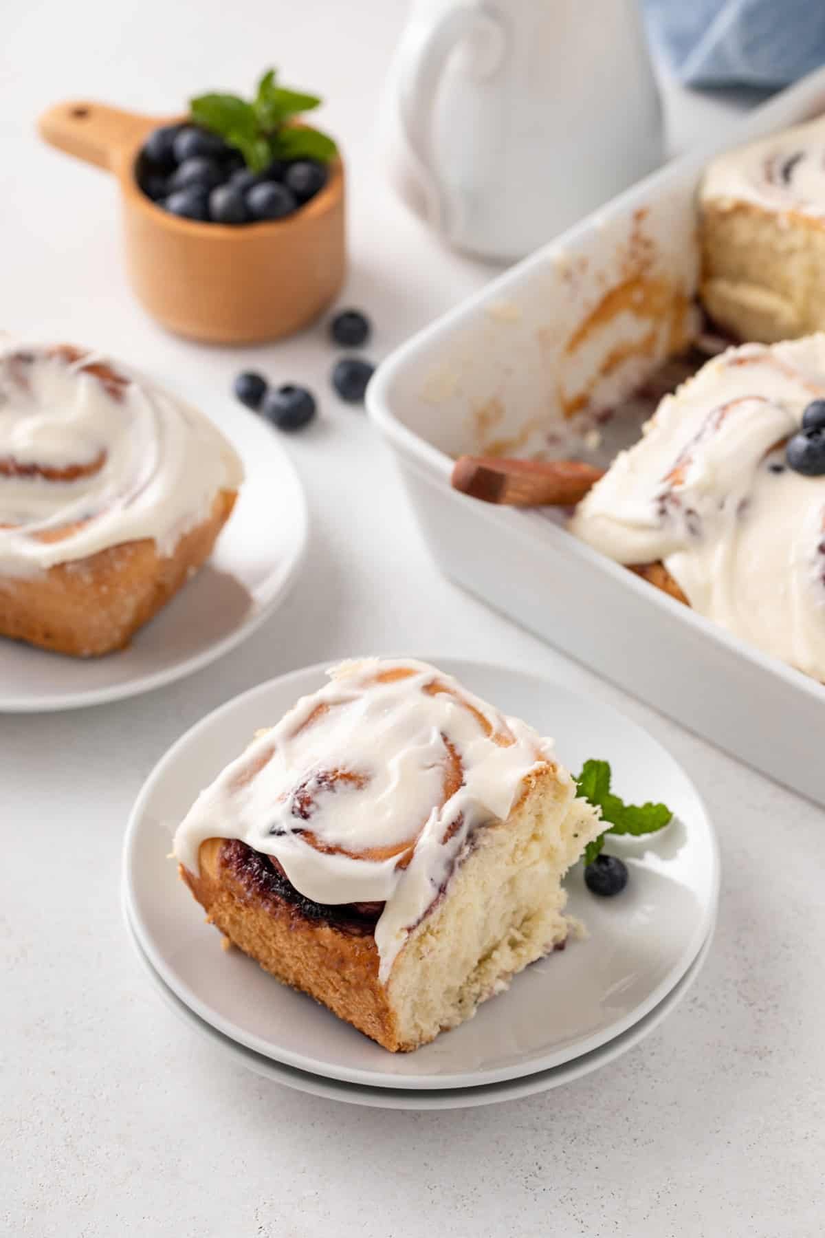 White plate holding a blueberry sweet roll, with a second plate and the pan of rolls in the background.