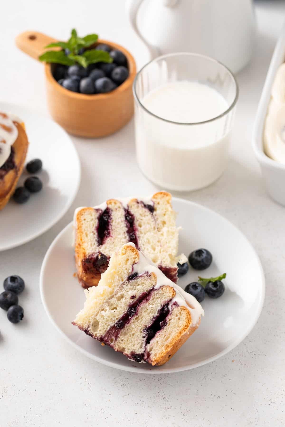 Blueberry sweet roll cut in half on a white plate in front of a glass of milk.