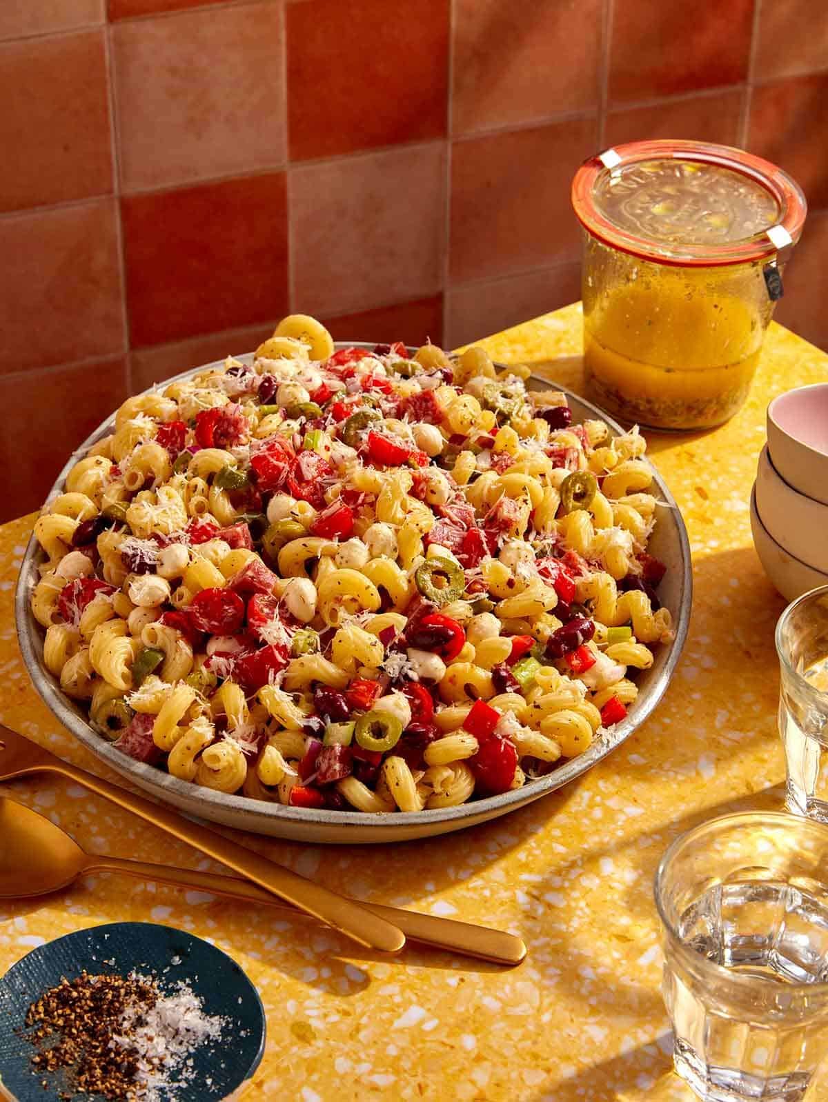 Pasta salad on a table with serving utensils and a jar of Italian vinaigrette.
