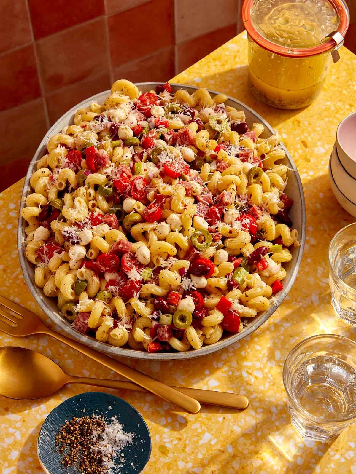 Pasta salad on a table with serving utensils and a jar of Italian vinaigrette.