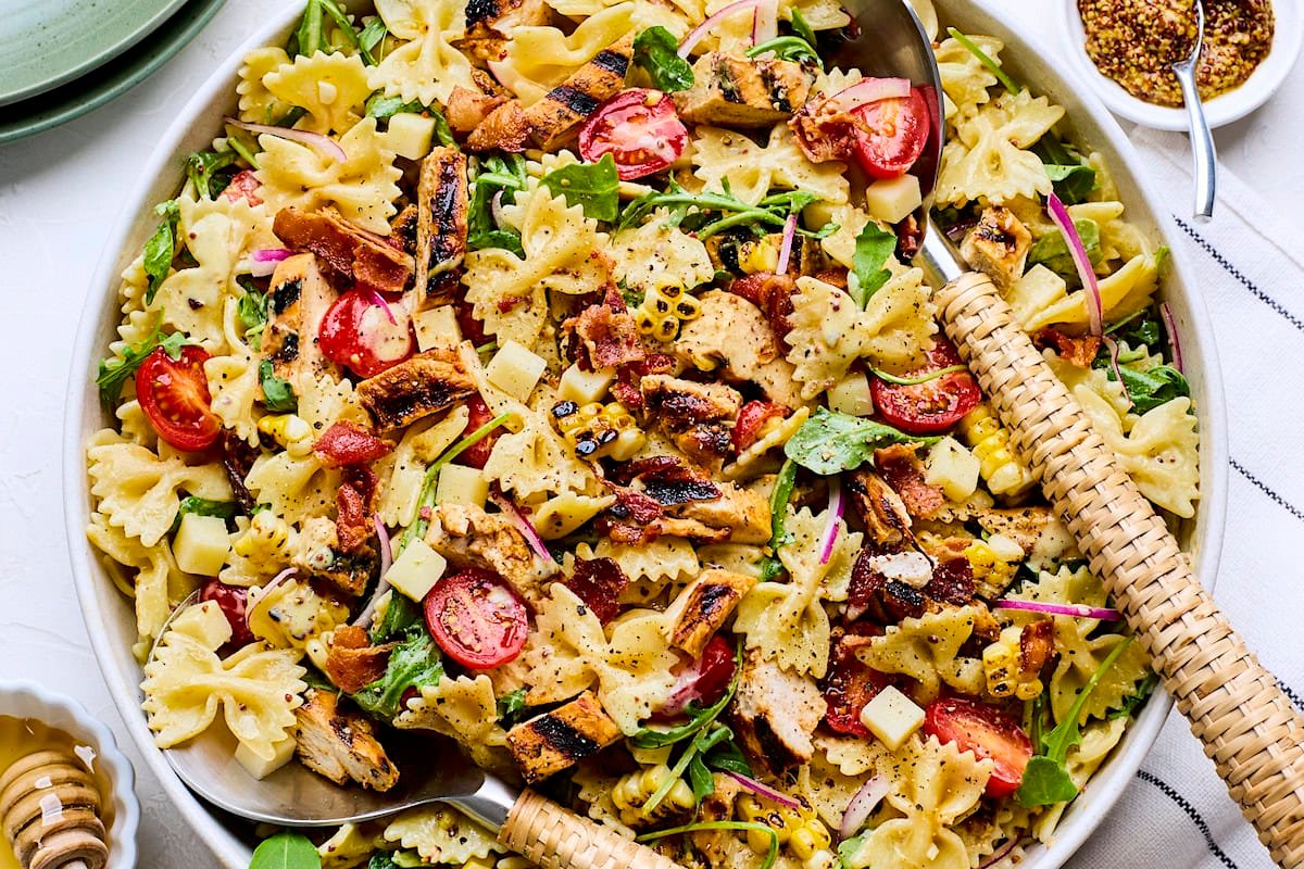 close up of honey mustard chicken pasta salad in serving bowl with serving utensils. 