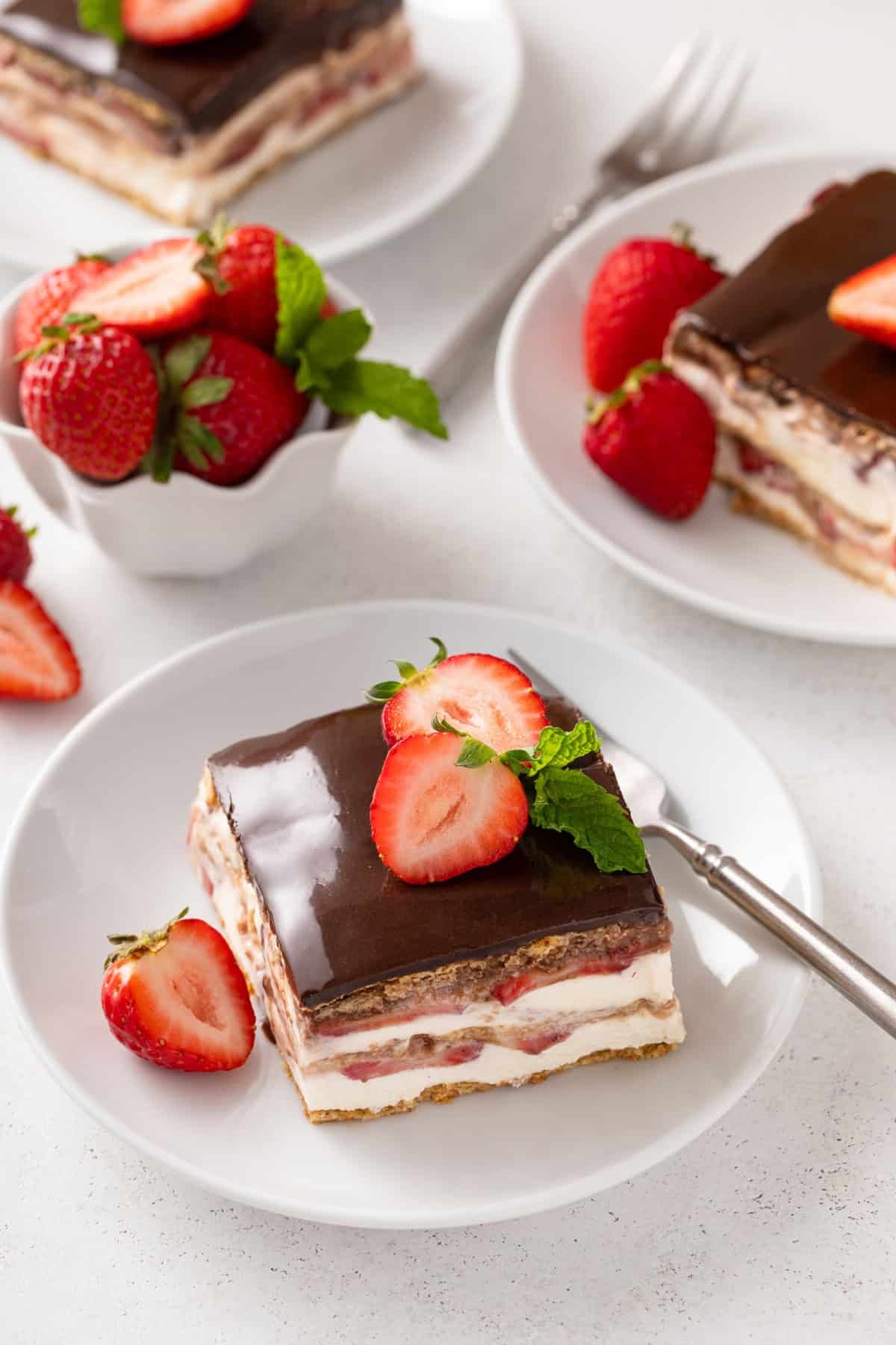 Plated slice of strawberry eclair cake on a countertop with two other plates and a bowl of fresh strawberries in the background.