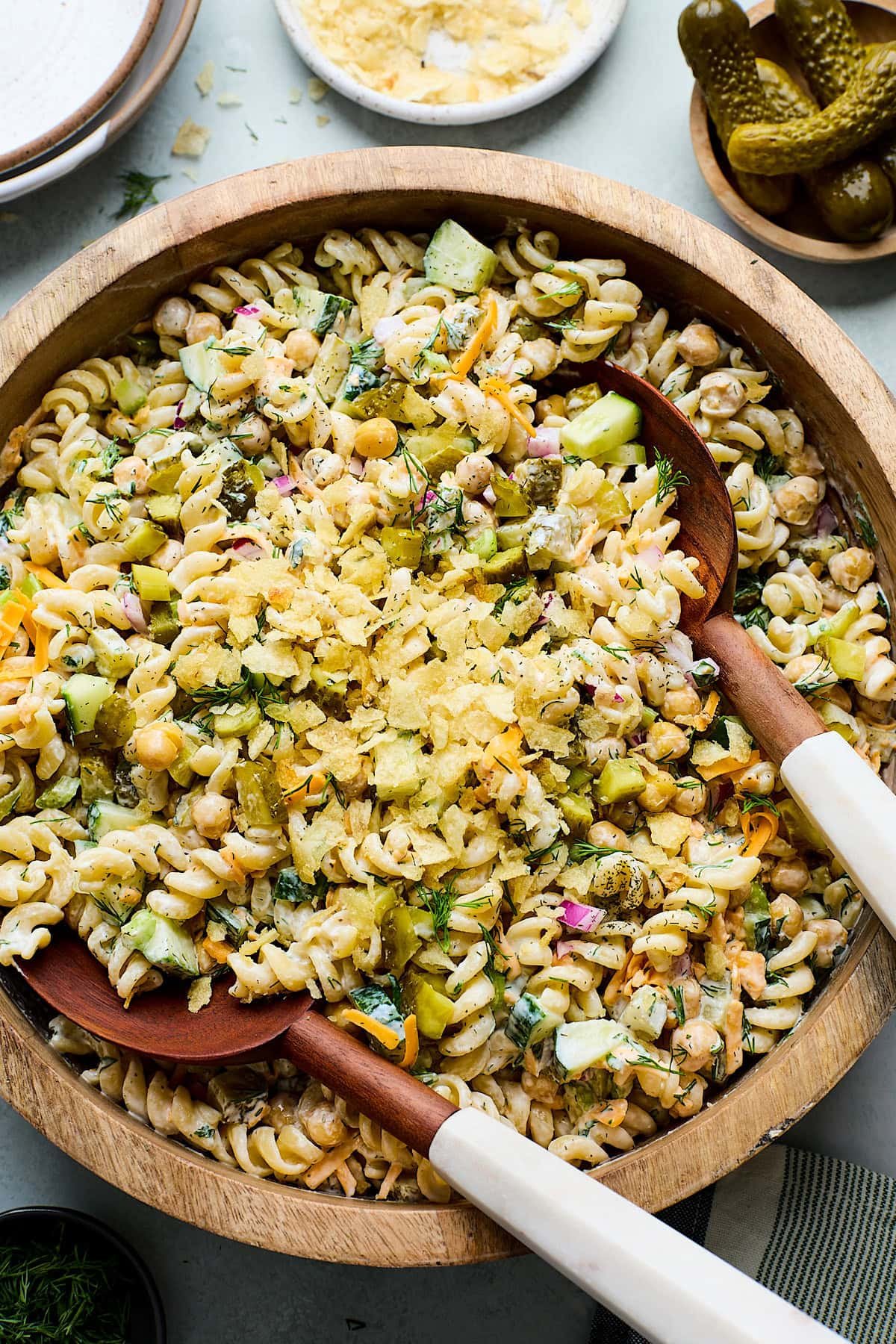 dill pickle pasta salad topped with crushed potato chips in bowl with salad servers. 