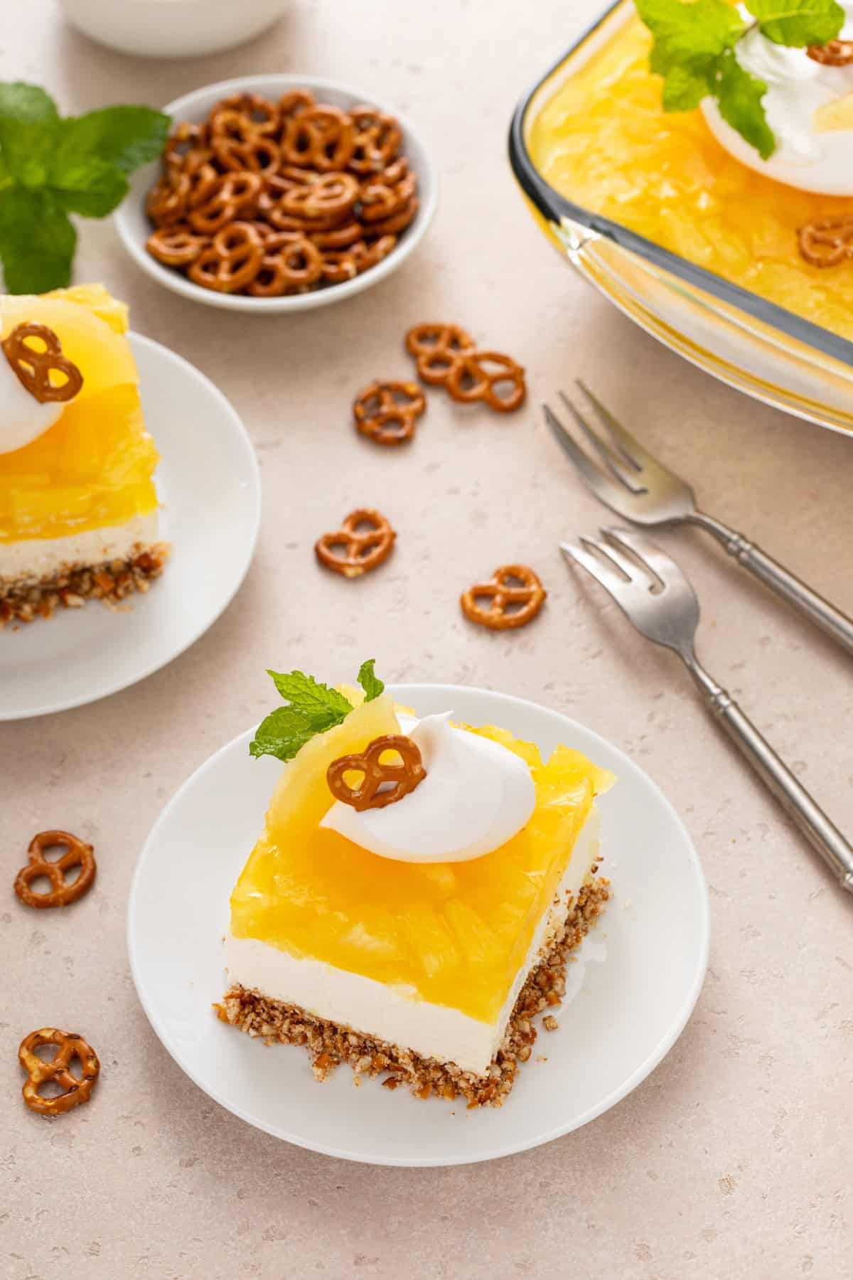 Two white plates holding slices of pineapple pretzel salad, with pretzel twists and a pan of the dessert in the background.