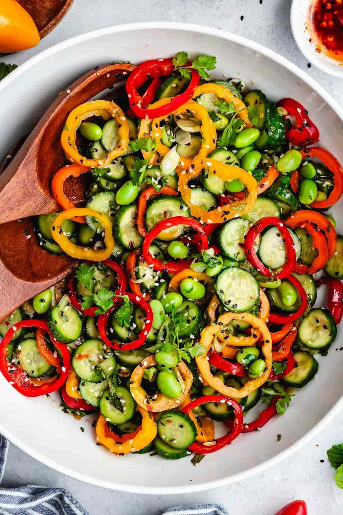 close up of cucumber sweet pepper salad with edamame in bowl with salad servers. 