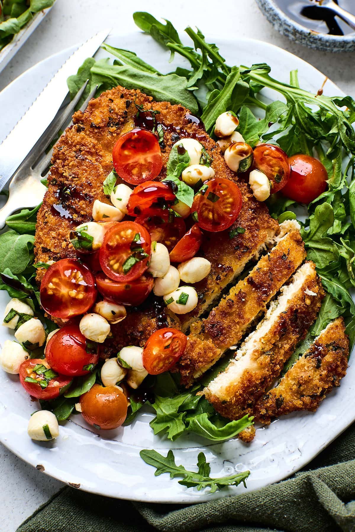 crispy chicken cutlet on plate with caprese salad on top with balsamic glaze and basil. 