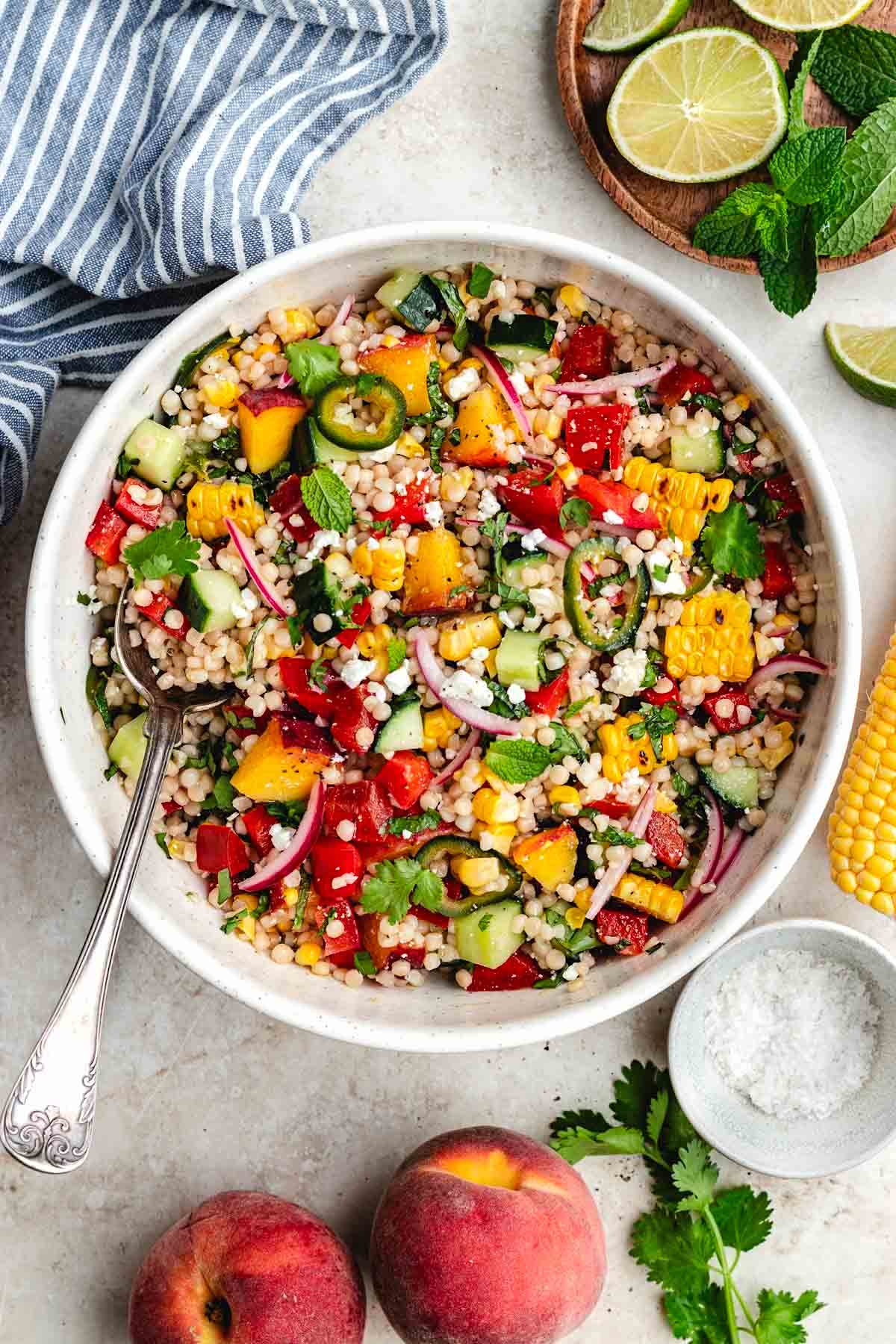 summer couscous salad with peaches, corn, cucumber, peppers, pickled red onion and jalapeño, with honey lime dressing in bowl with spoon.