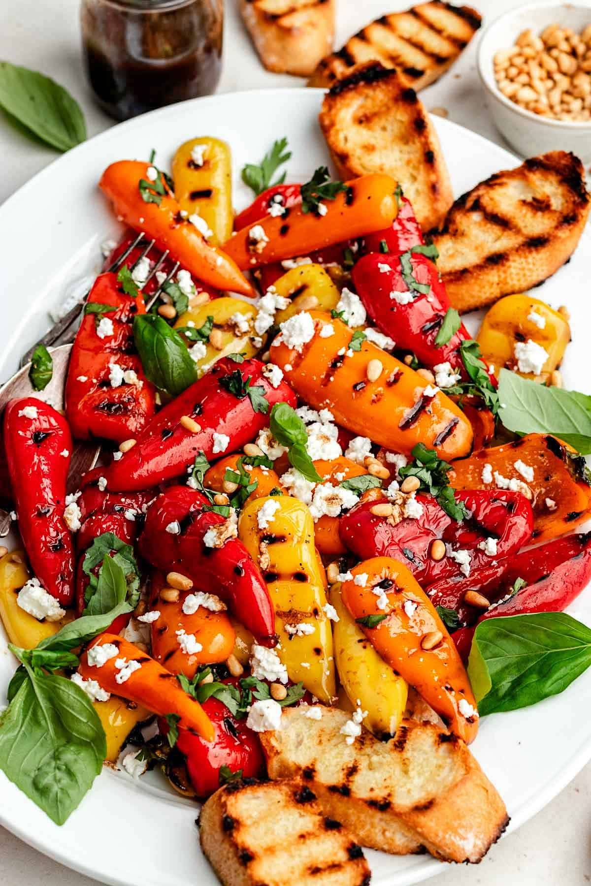 close up of grilled mini sweet peppers with basil, feta, pine nuts, and balsamic vinaigrette on platter with grilled bread. 