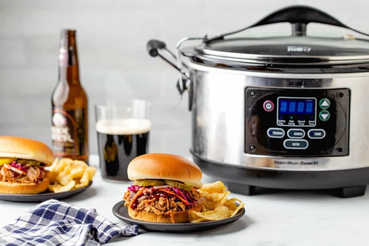 Pulled pork topped with slaw and pickles with a side of potato chips on a plate in front of a crock pot