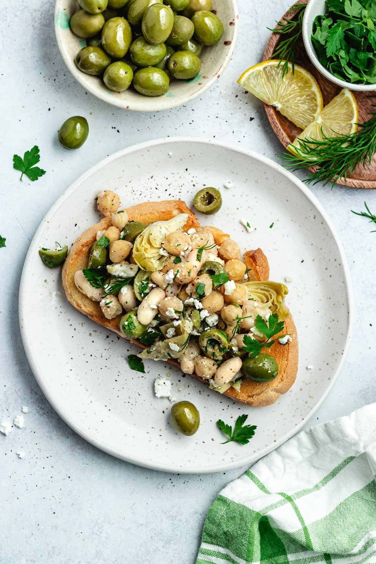 bean salad with artichokes, olives, feta, and herbs on toast on plate. 